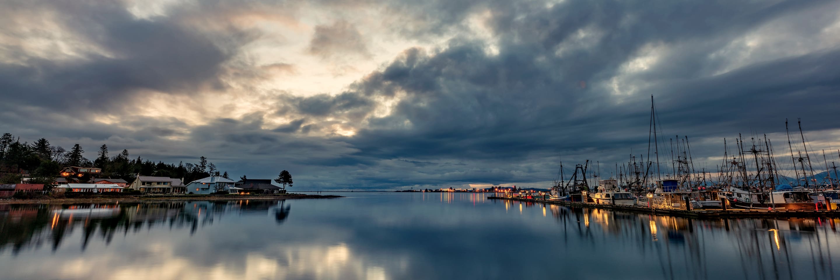 The fishermen's wharf at Comox at dusk, boats tied up under a heavy sky and lights on along the shore.