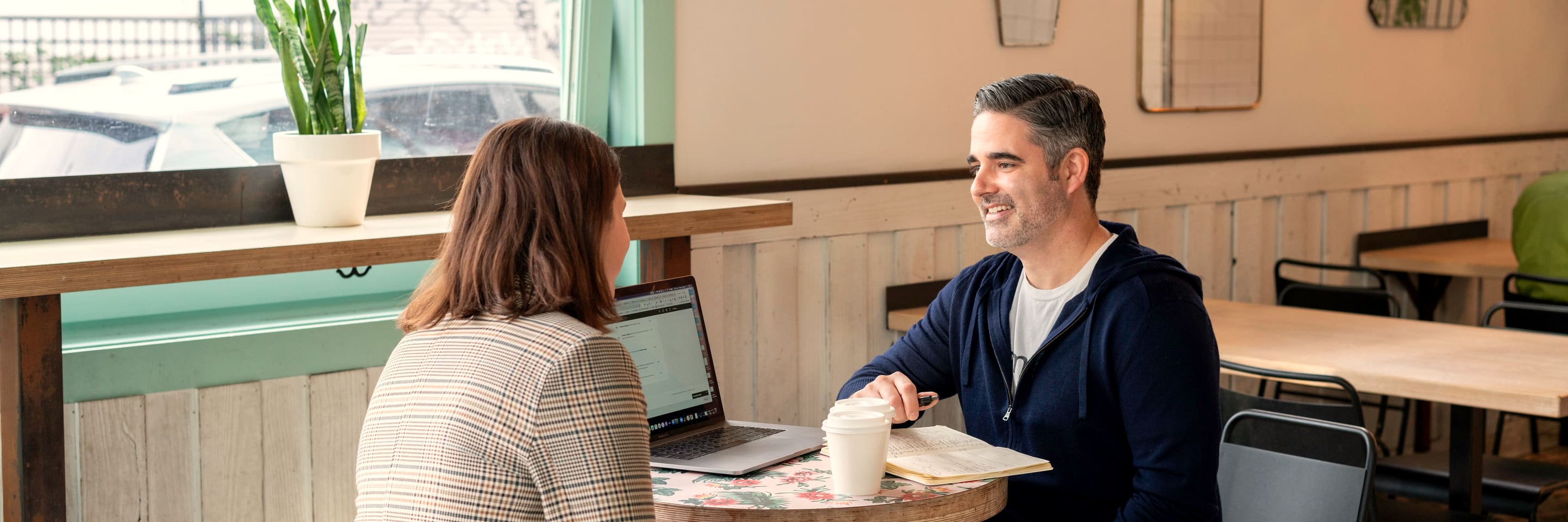 Two people in a one-to-one meeting at a cafe table by the window, a laptop open between them.