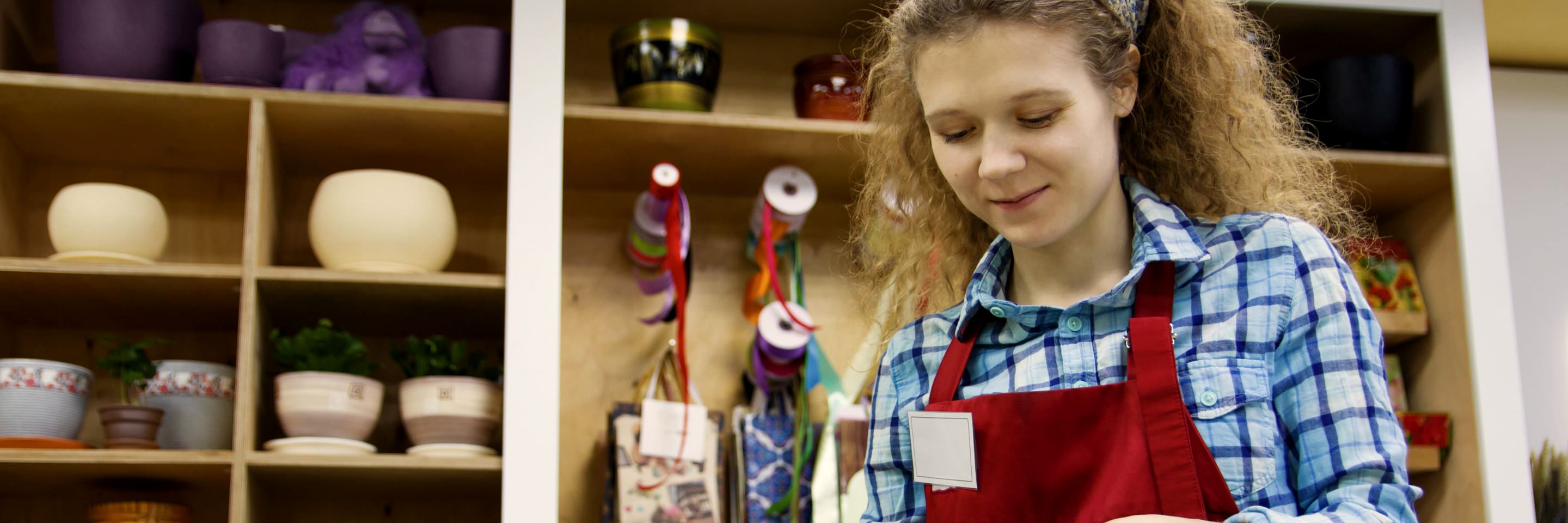 A florist in a red apron trimming roses at her counter, shelves of pots behind her.