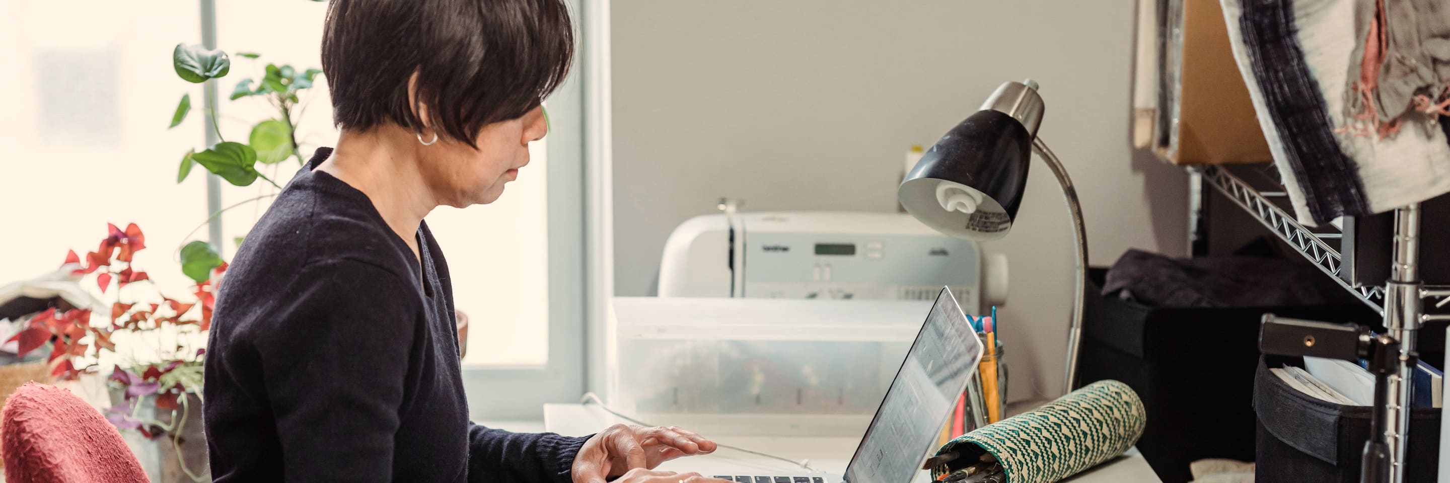 A woman typing at a laptop in her own workroom, a sewing machine and shelving behind her.