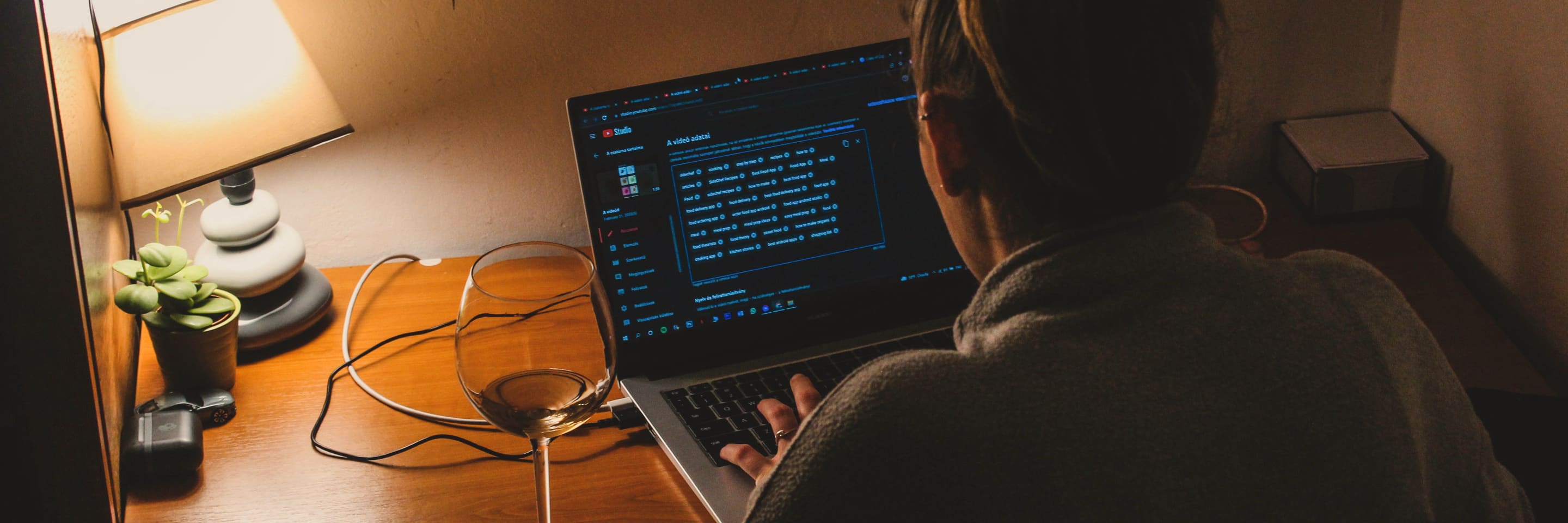 Someone working at a laptop late in the evening, a desk lamp and a glass of wine beside them.