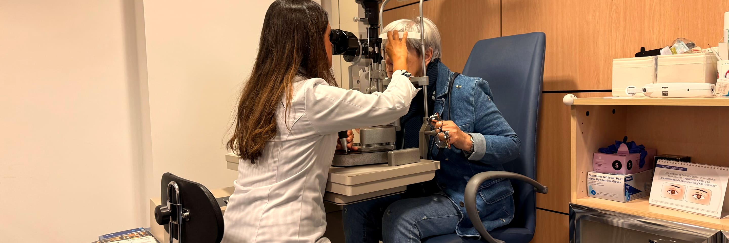 An optometrist in the middle of an eye exam, leaning into the slit lamp across from her patient.