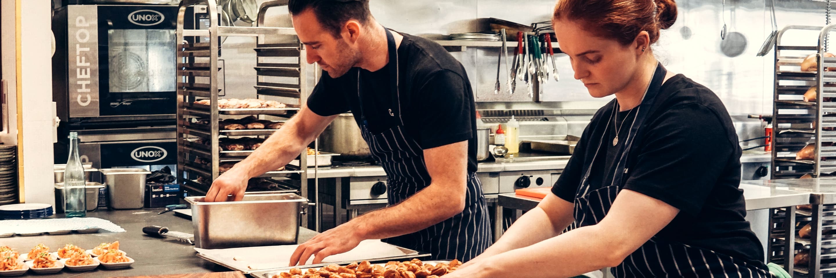 Two cooks plating at the steel counter of a restaurant kitchen, racks and combi oven behind them.