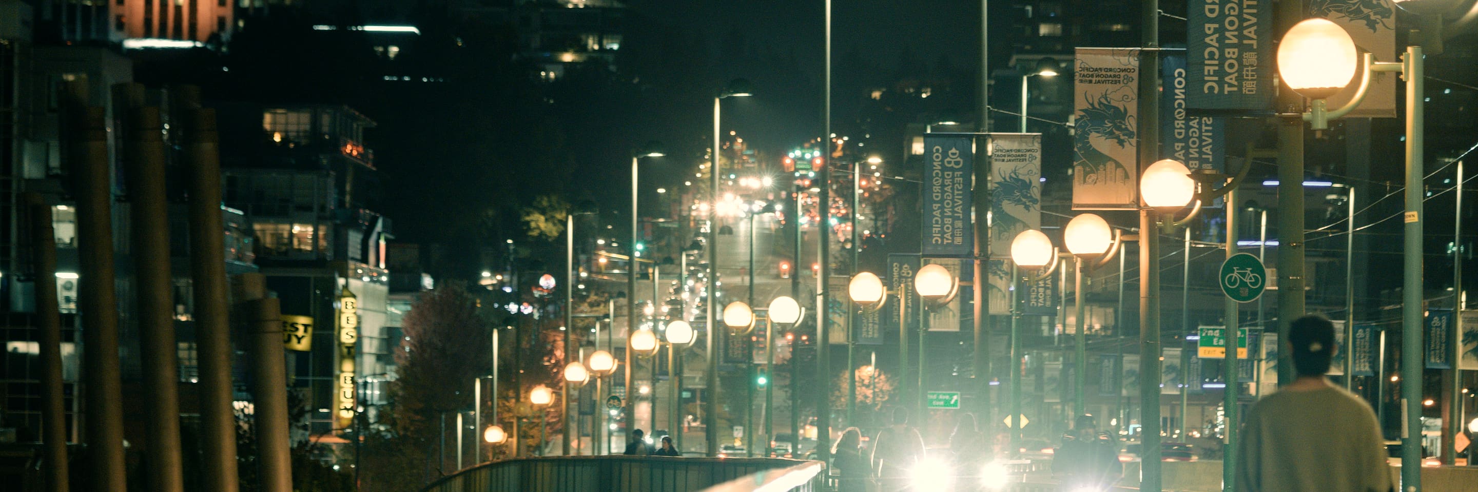 A Vancouver street at night, the lamp posts lit along the sidewalk and traffic coming up the hill.