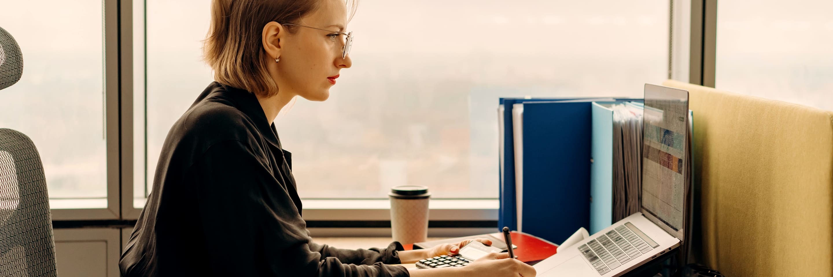 A bookkeeper at her desk by the window, calculator and laptop out, binders stacked beside her.