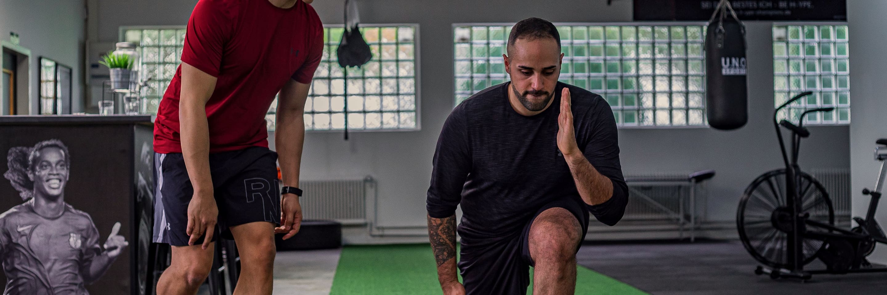 A trainer coaching a client through a lunge in a small gym with glass-block windows.