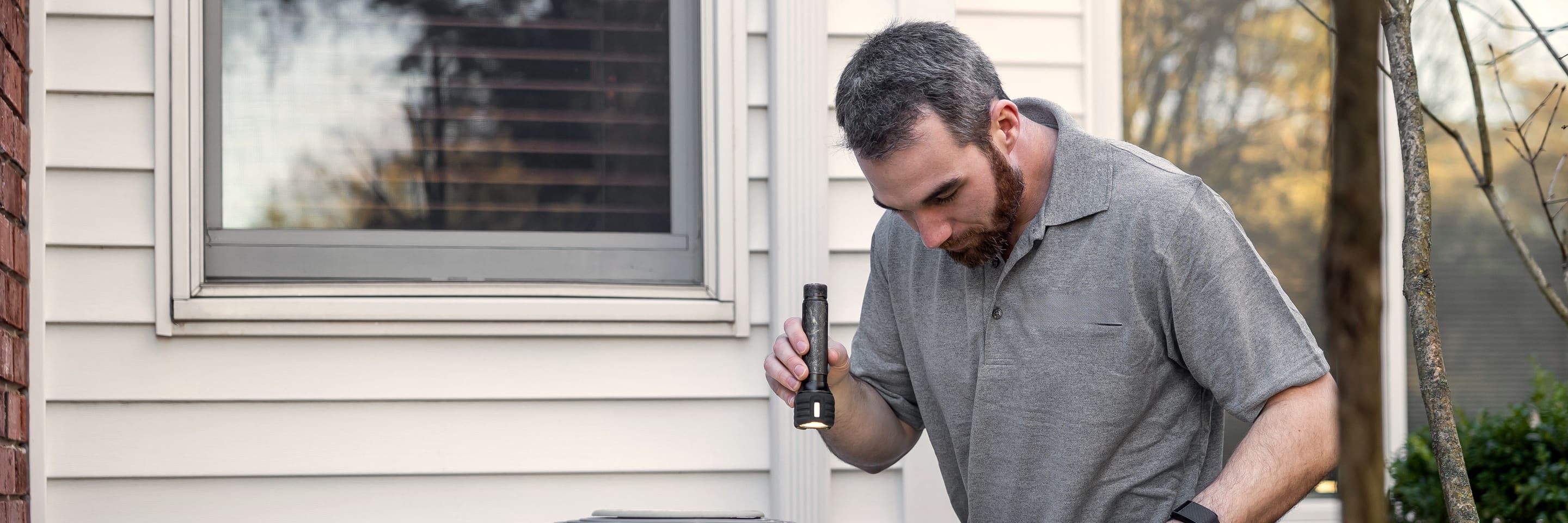 A technician with a flashlight inspecting the outdoor unit beside a house.