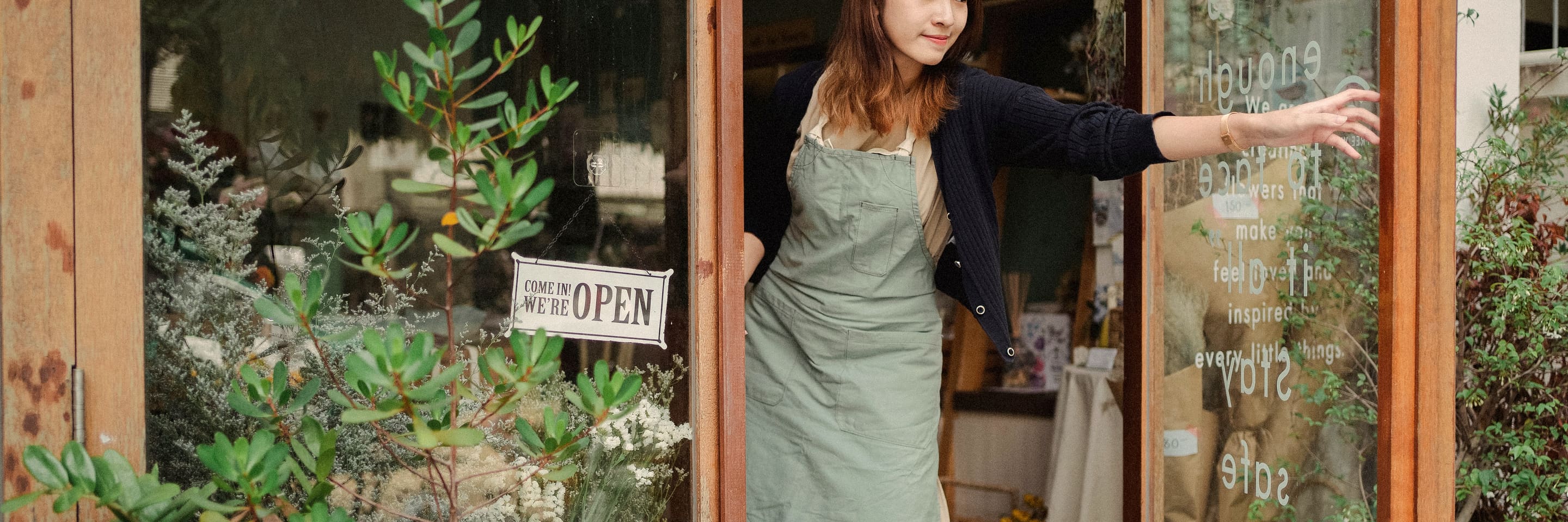 A shop owner in an apron opening the glass door of her store, a Come In We're Open sign in the window.