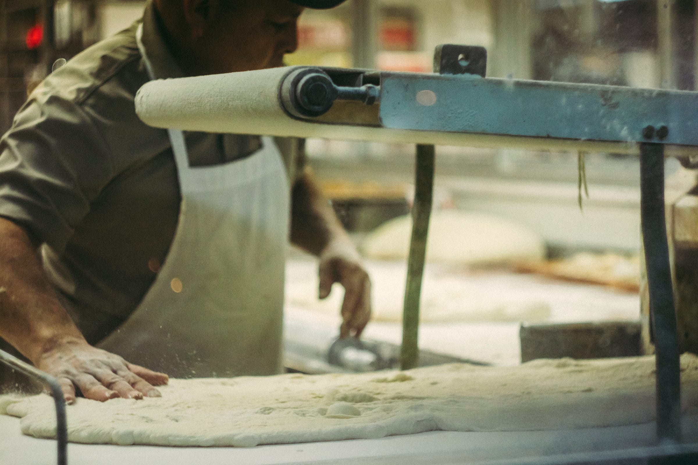 A baker in an apron working a sheet of dough through the rolling machine in a bakery.
