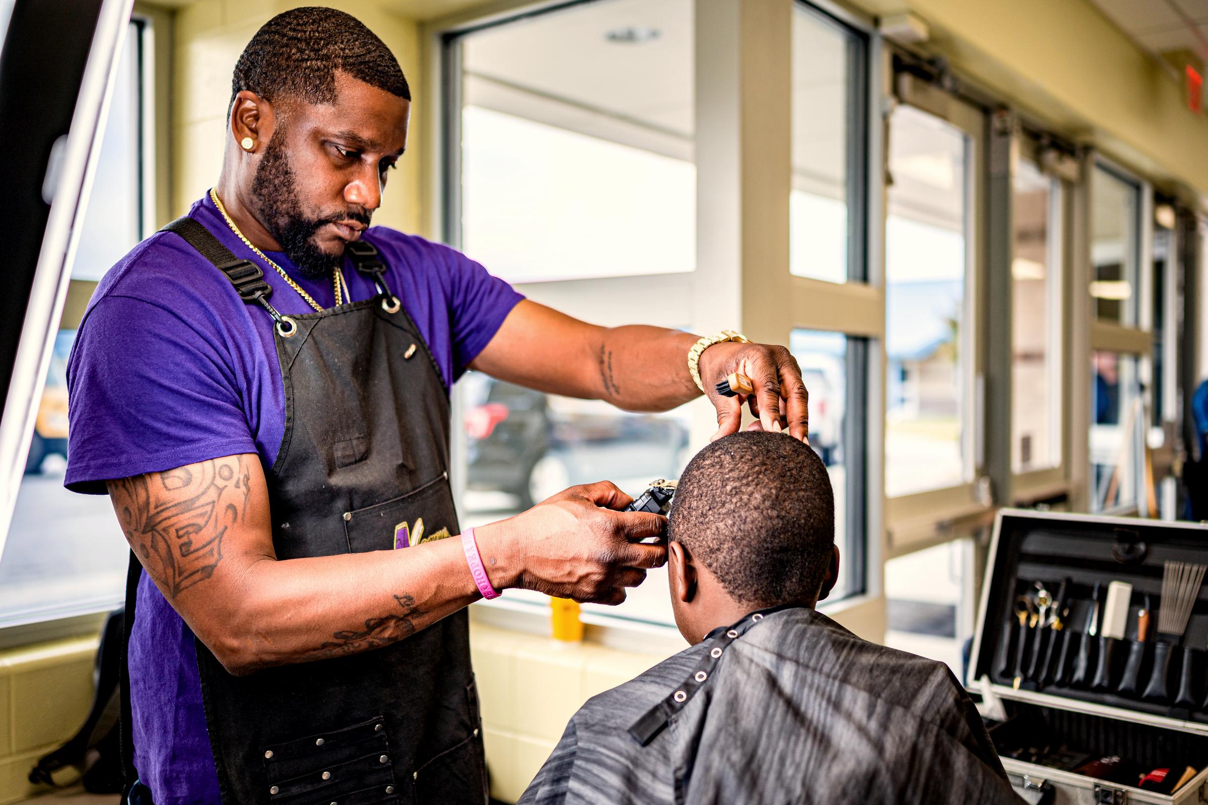 A barber in an apron running clippers over a seated client's head in a bright shopfront, his kit open on the counter.