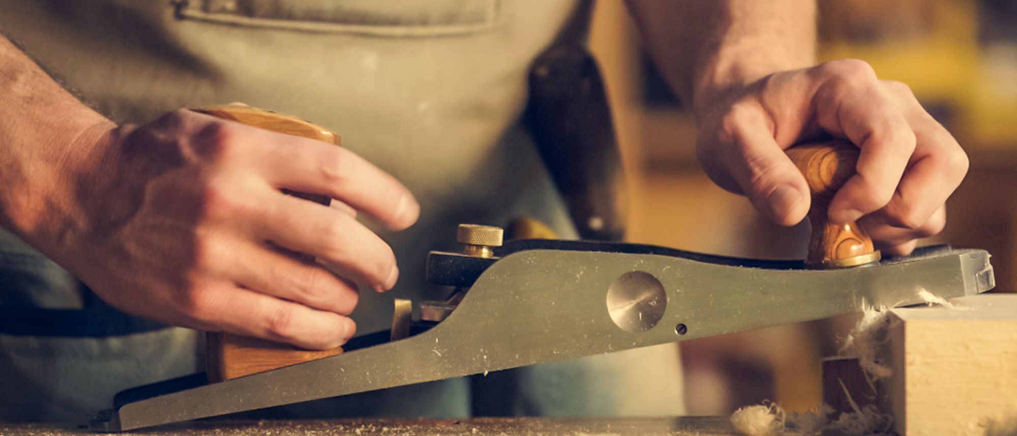 Hands guiding a hand plane along a board in a warmly lit workshop.