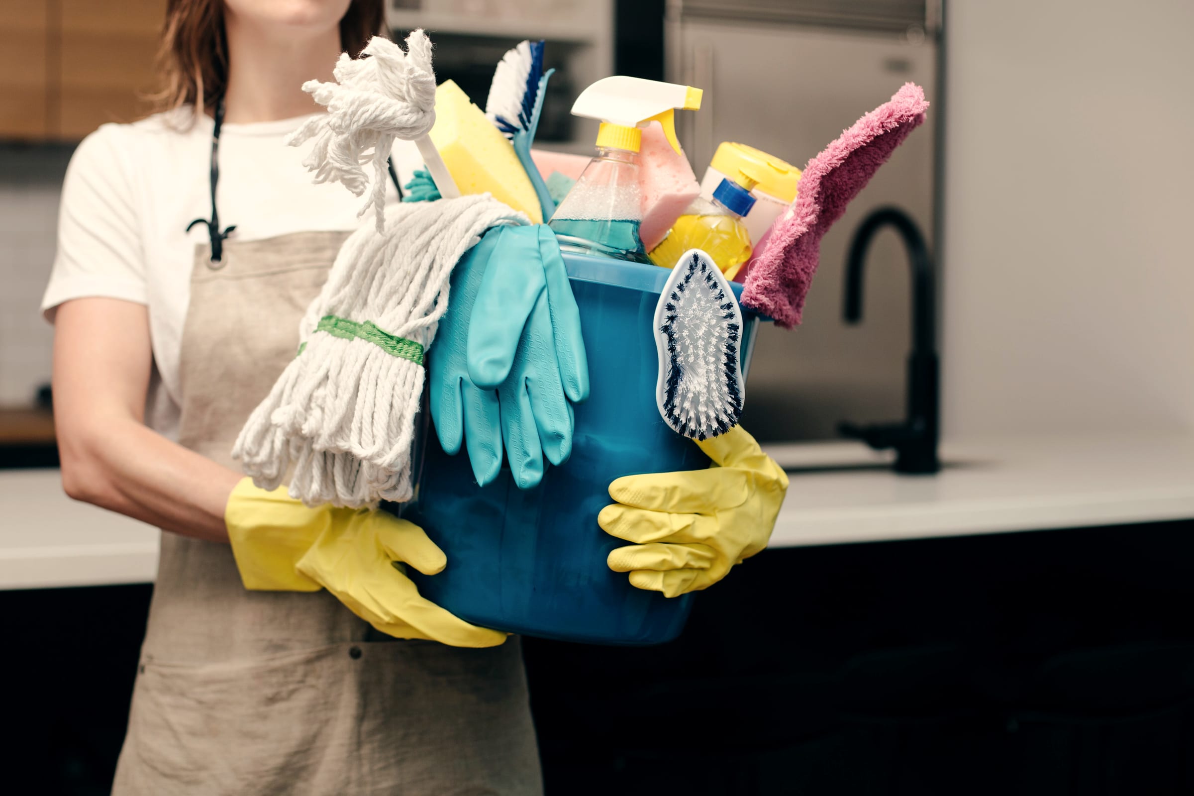 A cleaner in an apron carrying a bucket of supplies, mop and gloves through a kitchen.