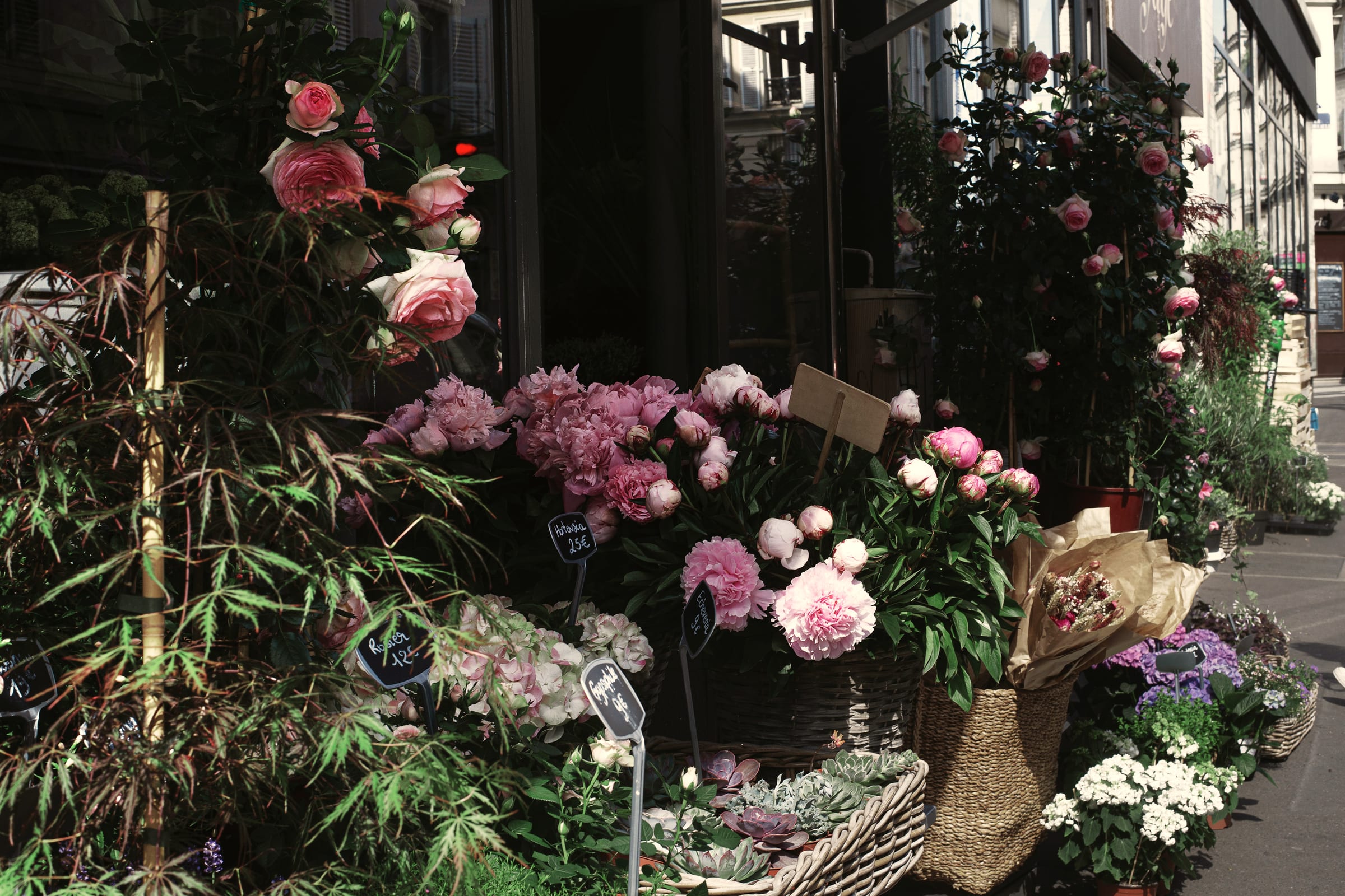 The front of a flower shop, buckets of roses and peonies banked along the pavement.