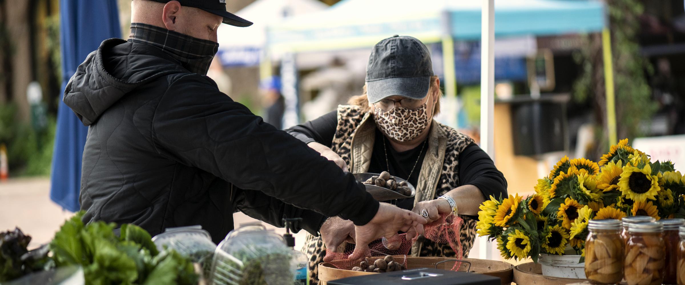 A stallholder handing produce across the table to a customer at an outdoor market, sunflowers and preserves beside her.