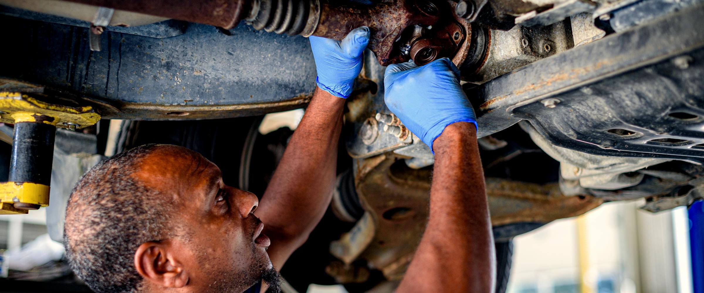 A mechanic on his back under a raised vehicle, gloved hands working at a driveshaft joint.
