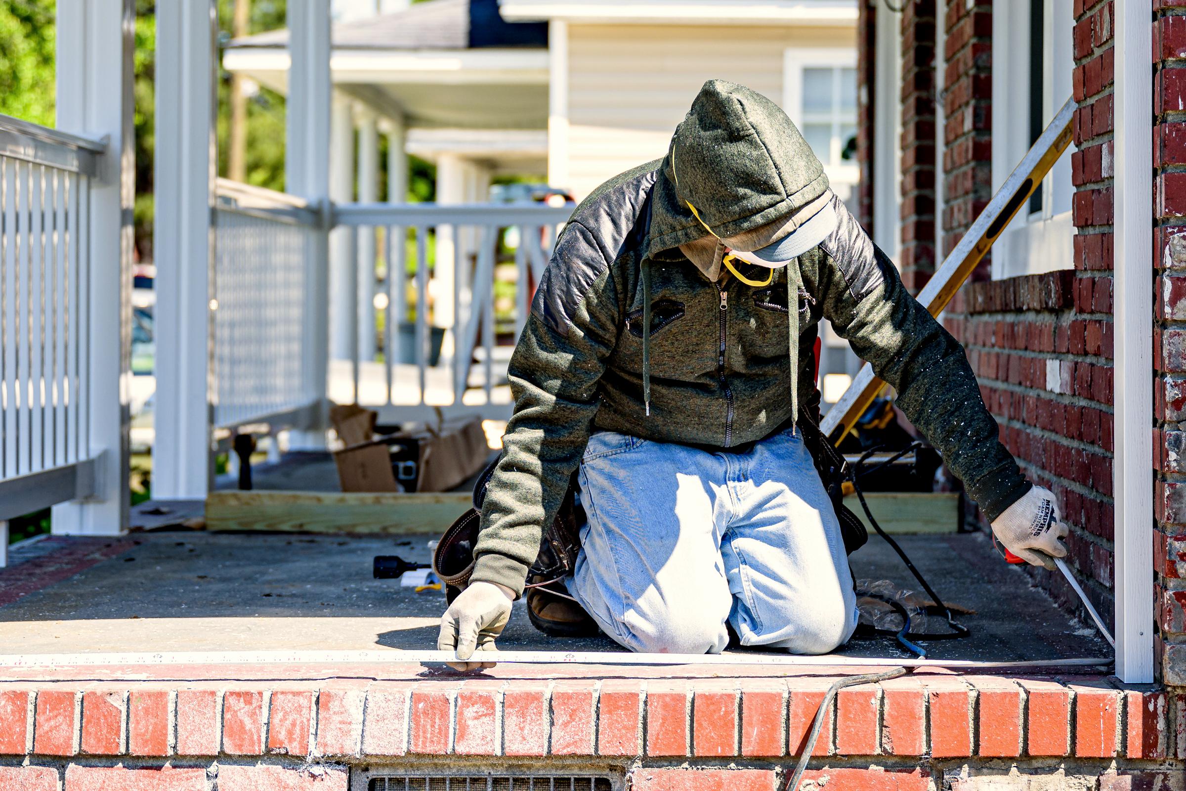 A painter in overalls and a hood kneeling on a brick porch, cutting in the edge of a repainted house front.