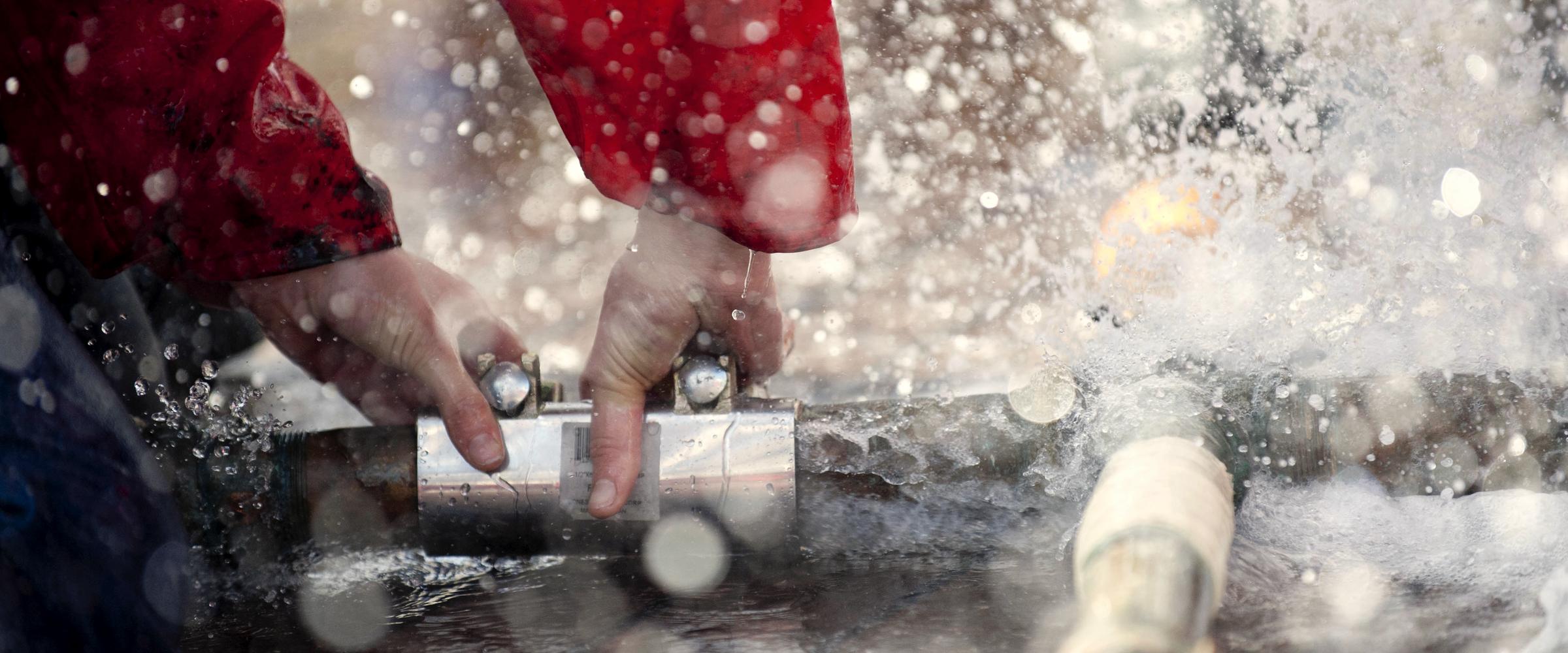 Two pairs of hands joining a length of pipe with a repair coupling while water sprays across the frame.