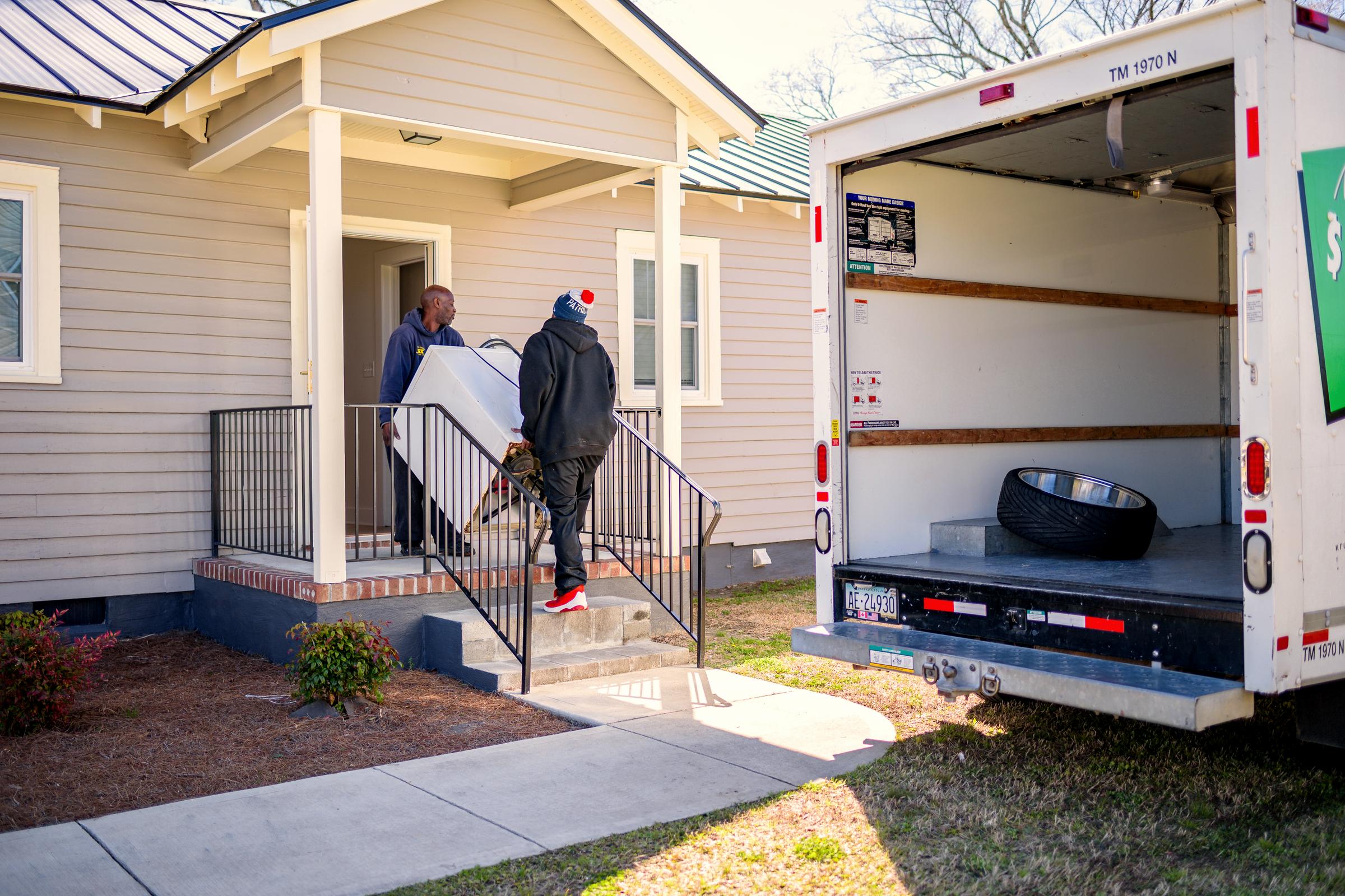 Two people carrying furniture up the steps of a small clapboard house, a hired box truck open at the kerb.