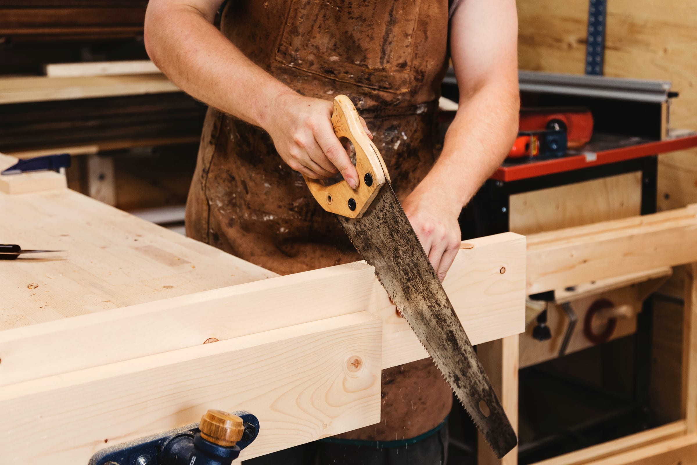 A woodworker in a leather apron cutting a board with a handsaw at the bench.